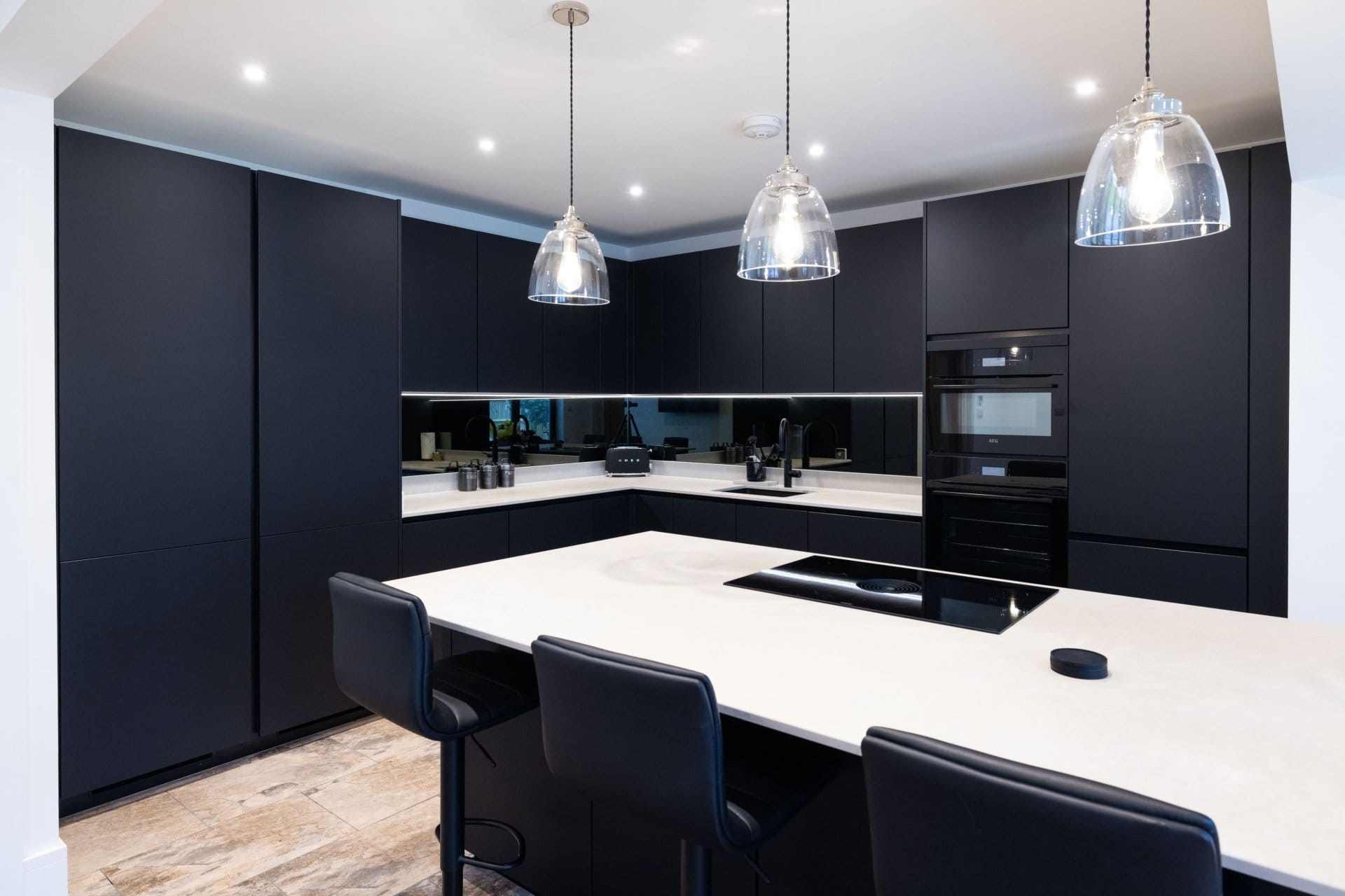 Artizan Interiors modern dark handleless kitchen featuring matte black cabinetry, a white island breakfast bar, integrated appliances and contemporary pendant lighting.